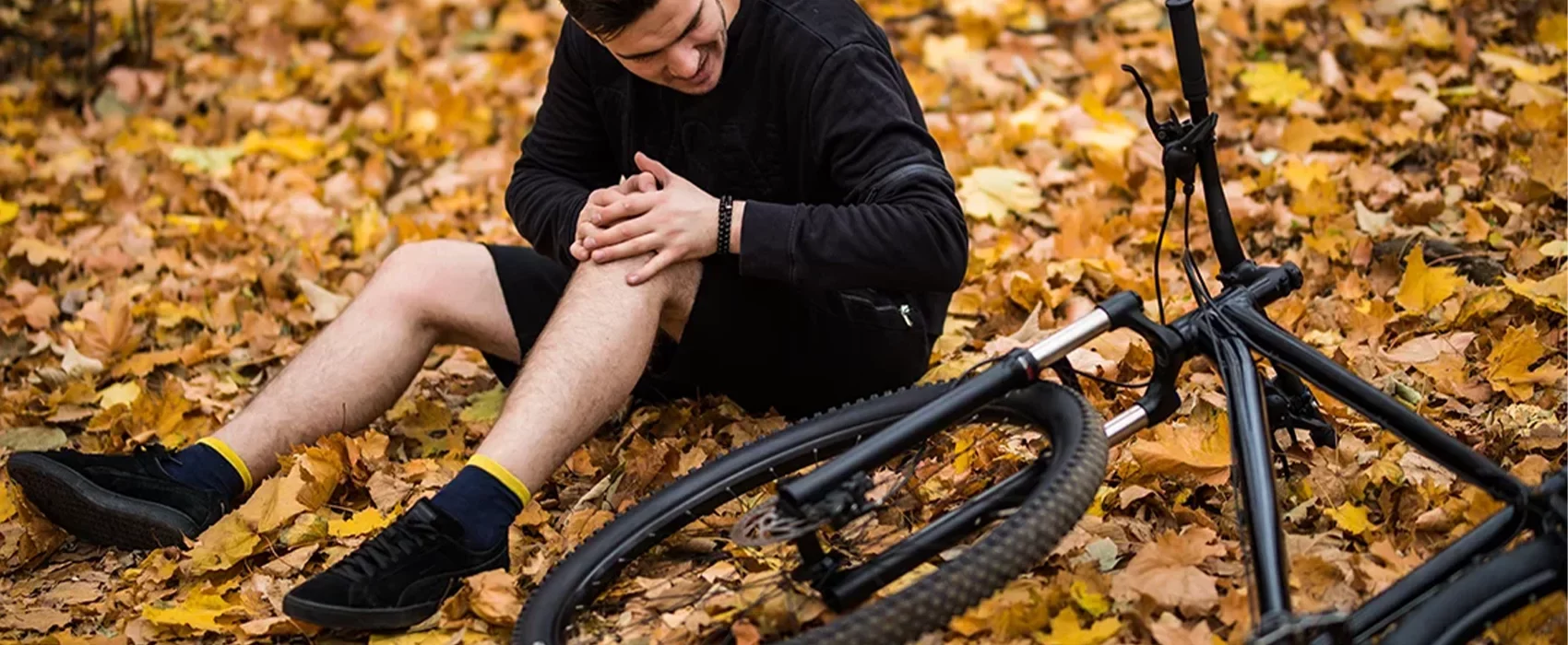 active young man holding by his hurt broken leg while lying autumn forest path by his bicycle